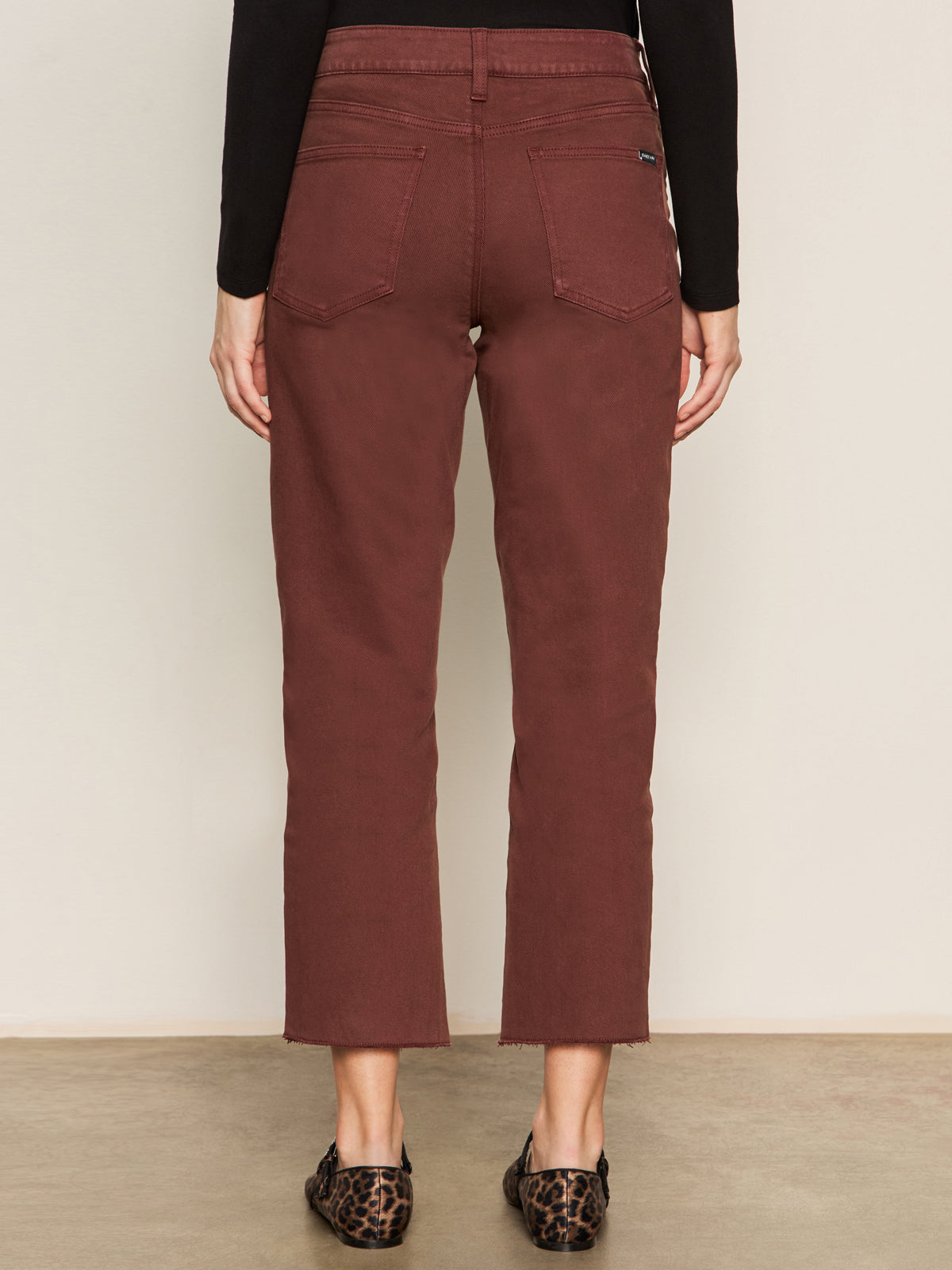 A person wears Sanctuary Clothing’s cadence 90s straight crop jeans in chicory, featuring a cropped, straight-leg maroon style with raw hems and back pockets. They pair it with a black long-sleeve top and leopard-print flats.