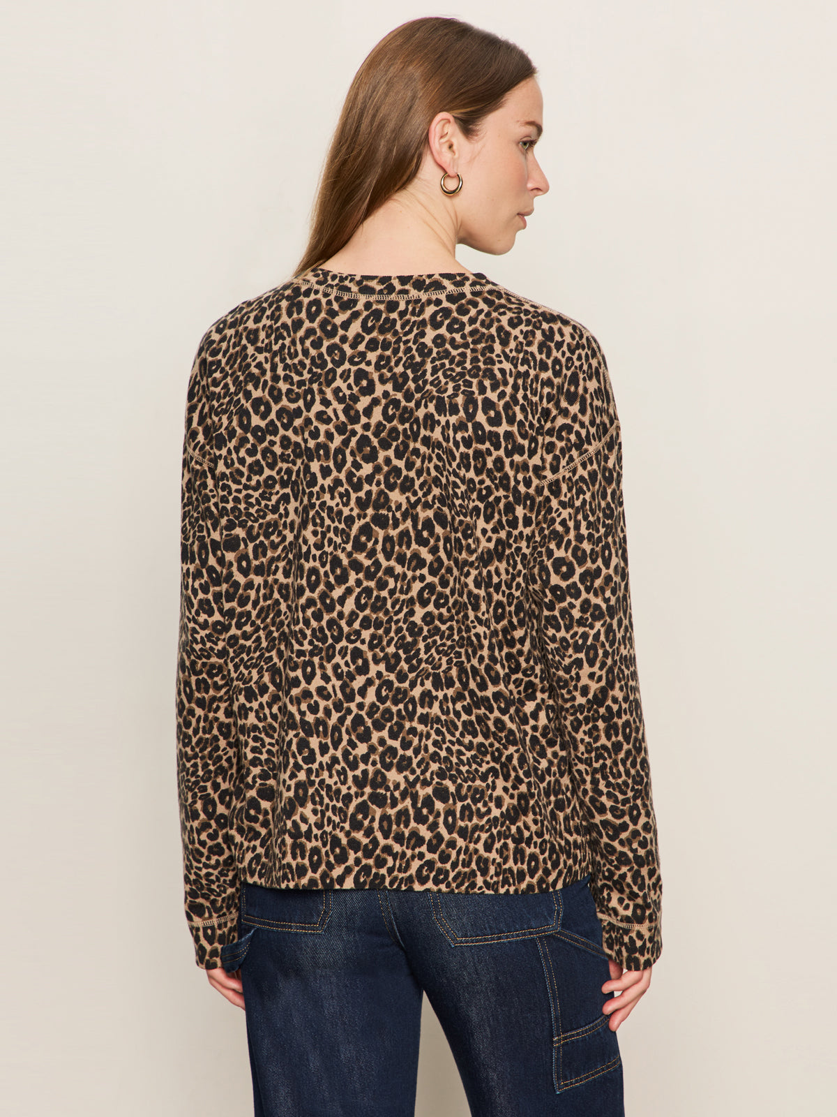 A woman with long brown hair is seen from behind, wearing the Sanctuary Clothing 24 7 Popover French Kitty top and dark blue jeans, standing against a plain light-colored background.
