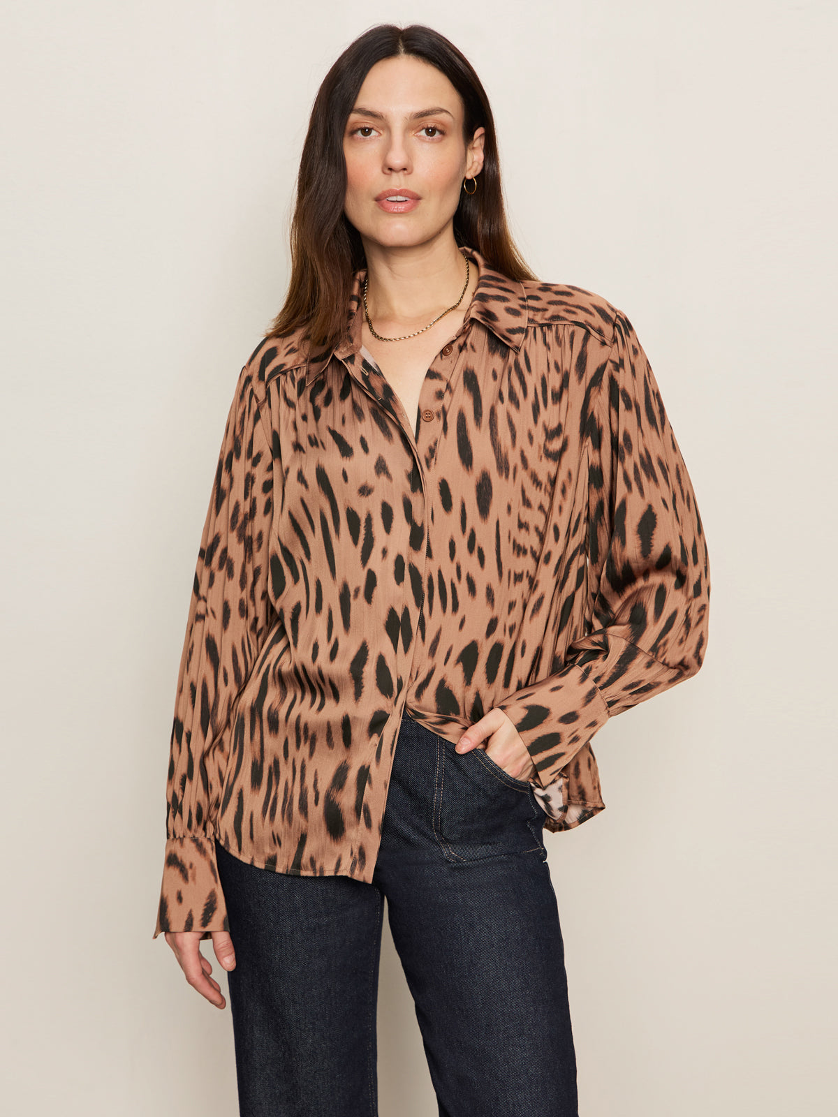 A woman with long brown hair wears the Sanctuary Clothing Colette Shirt in mocha leo, a loose-fitting leopard print blouse, paired with dark jeans. She stands against a plain light-colored background, one hand in her pocket, looking at the camera.
