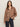 A woman with long brown hair wears the Sanctuary Clothing Colette Shirt in mocha leo, a loose-fitting leopard print blouse, paired with dark jeans. She stands against a plain light-colored background, one hand in her pocket, looking at the camera.