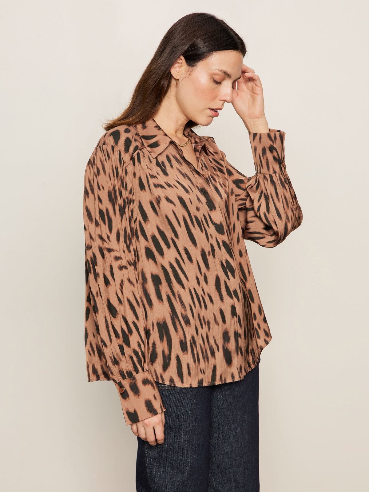 A woman with straight brown hair wears the Sanctuary Clothing colette shirt in mocha leo—a loose, long-sleeved, animal-print blouse with black and tan spots—and dark pants, facing sideways against a plain light background.