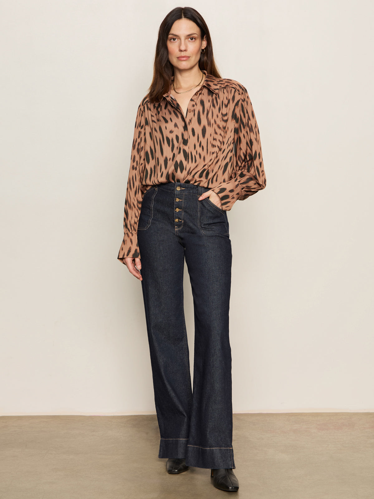 A woman with long, straight hair stands against a beige wall, wearing the Sanctuary Clothing Colette Shirt in mocha leo tucked into high-waisted, wide-leg dark denim jeans with buttons and front pockets. She completes the look with black shoes.