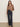 A woman with long, straight hair stands against a beige wall, wearing the Sanctuary Clothing Colette Shirt in mocha leo tucked into high-waisted, wide-leg dark denim jeans with buttons and front pockets. She completes the look with black shoes.