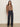 A woman with long hair stands against a plain wall wearing the Sanctuary Clothing ekara slim wide jean in indigo dark rinse wash with front buttons, paired with a brown animal-print blouse and black shoes.