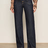 A person wears Sanctuary Clothing’s ekara slim wide jean in indigo dark rinse wash, featuring prominent seams and front buttons, paired with an animal print long-sleeve blouse and black shoes against a plain background.