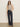 A woman stands against a light wall with one hand in her pocket, wearing Sanctuary Clothing’s fuzzy leopard shell mother leo top, sunglasses, high-waisted dark blue wide-leg jeans with buttons, and brown shoes.