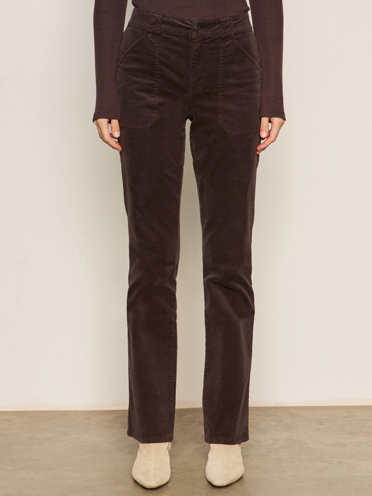 A person wears Sanctuary Clothing's Corduroy Hayden Bootcut Pant in Semi Sweet, featuring front patch pockets, styled with a dark long-sleeve top and light beige shoes, against a plain light backdrop.