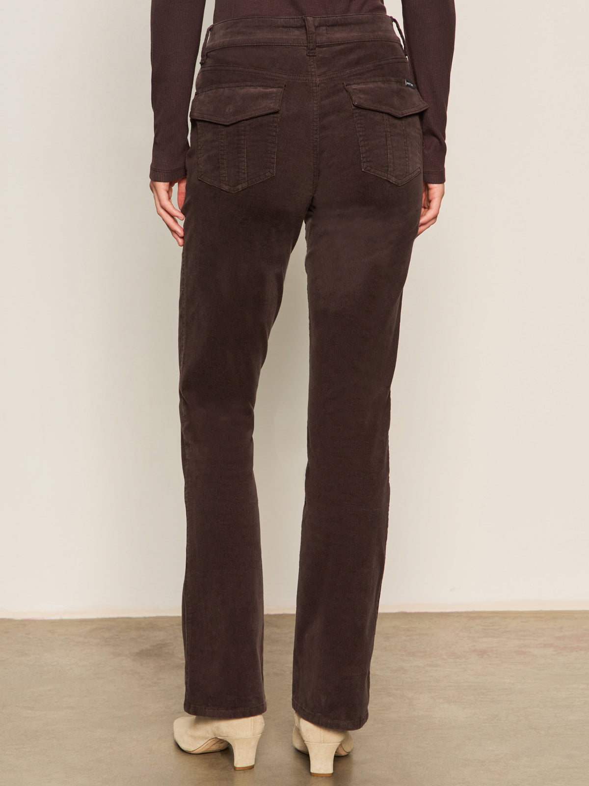 A person faces away wearing Sanctuary Clothing's corduroy Hayden bootcut pant in semi sweet, paired with a dark long-sleeve top and beige shoes. They stand on a light floor against a plain light wall.