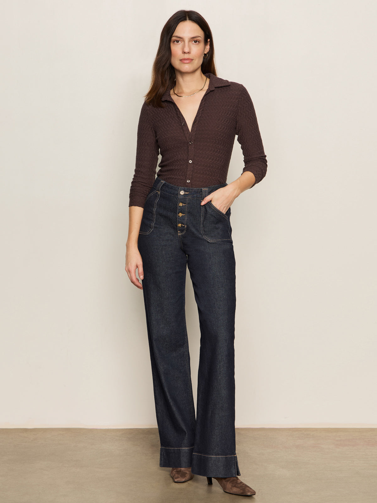 A woman stands against a plain light wall, wearing the Sanctuary Clothing textured knit dreamgirl top semi sweet with high-waisted dark blue wide-leg jeans and brown heeled shoes.