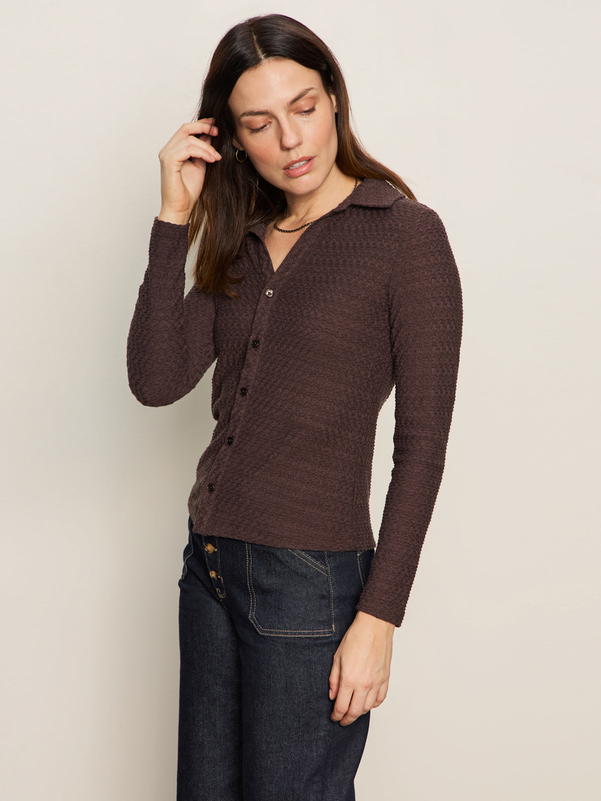 A woman with long brown hair wears the Sanctuary Clothing textured knit dreamgirl top semi sweet, a brown button-up with long sleeves, and dark high-waisted jeans. She stands against a light beige background, looking down and touching her hair.
