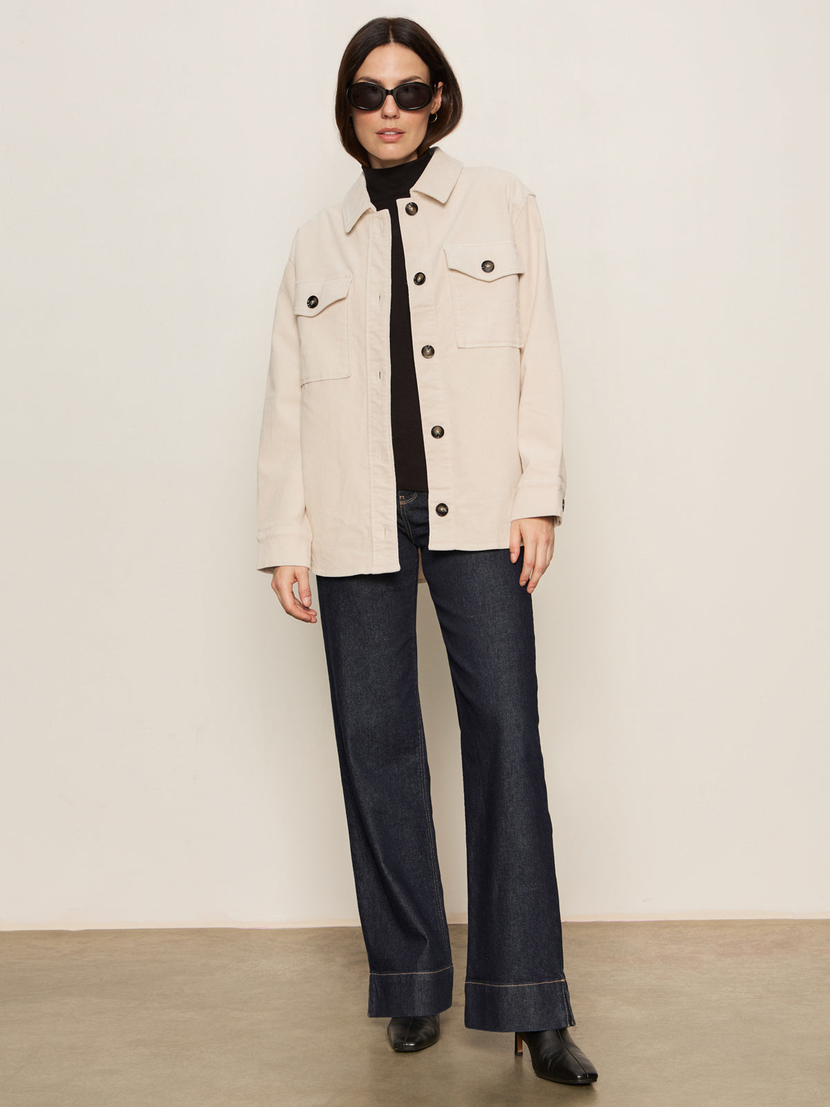 A woman wears Sanctuary Clothing's "the corduroy shacket mother of pearl" over a black top, paired with sunglasses, dark wide-leg jeans, and black heeled boots, standing against a plain light background.