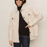 A woman stands against a plain background with one hand in her pocket, wearing the corduroy shacket mother of pearl by Sanctuary Clothing over a black turtleneck, paired with dark jeans and large black sunglasses.