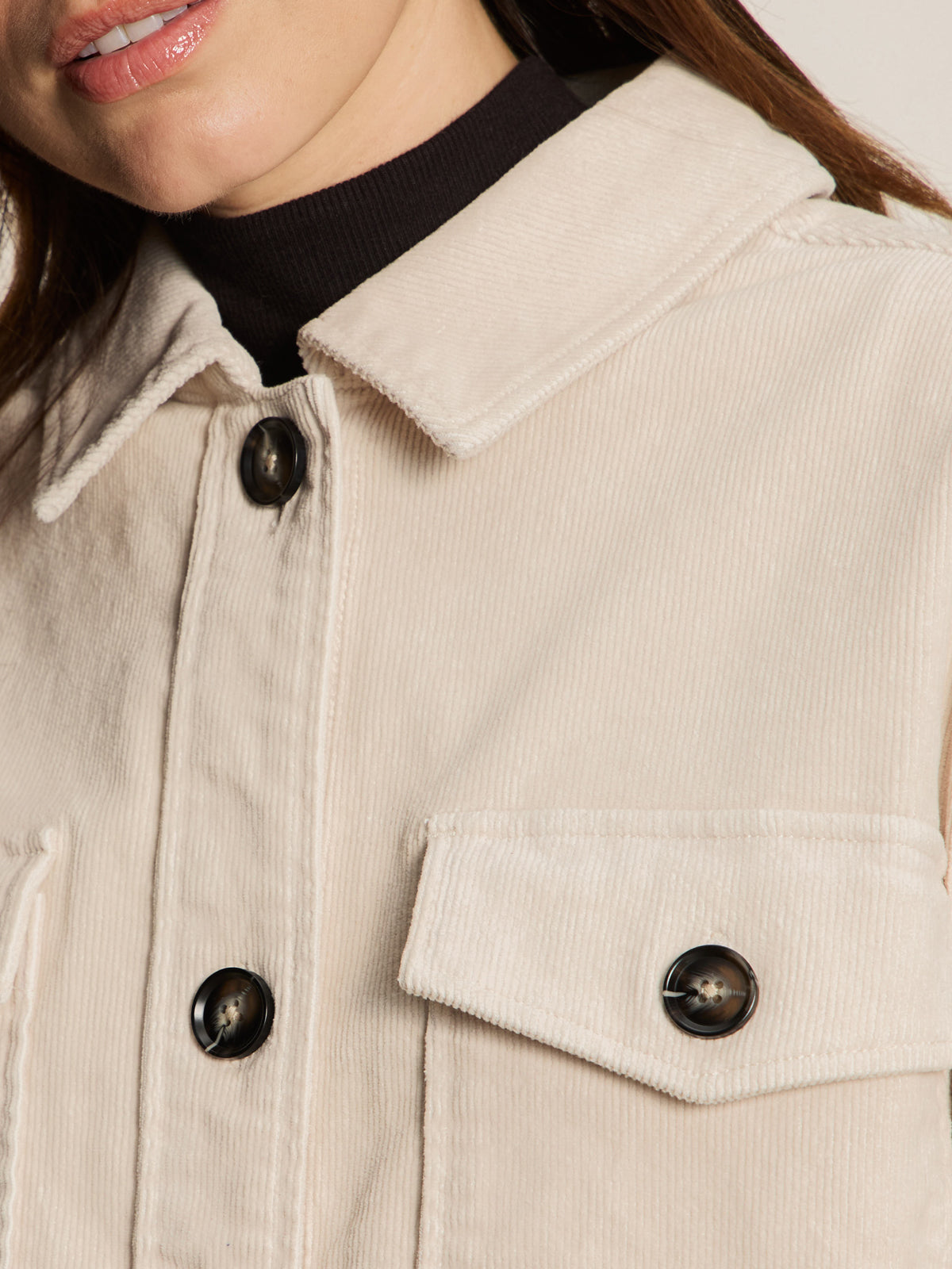 Close-up of a person wearing Sanctuary Clothing’s “the corduroy shacket mother of pearl”—a light beige jacket with black buttons and front pocket—over a black turtleneck, showing only the lower half of the face and upper torso.