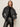 Wearing the Sanctuary Clothing boyfriend trucker jacket in washed black denim, a person stands against a plain light background with hands in pockets, paired with a leopard print collar, black knit sweater, and black leather pants.