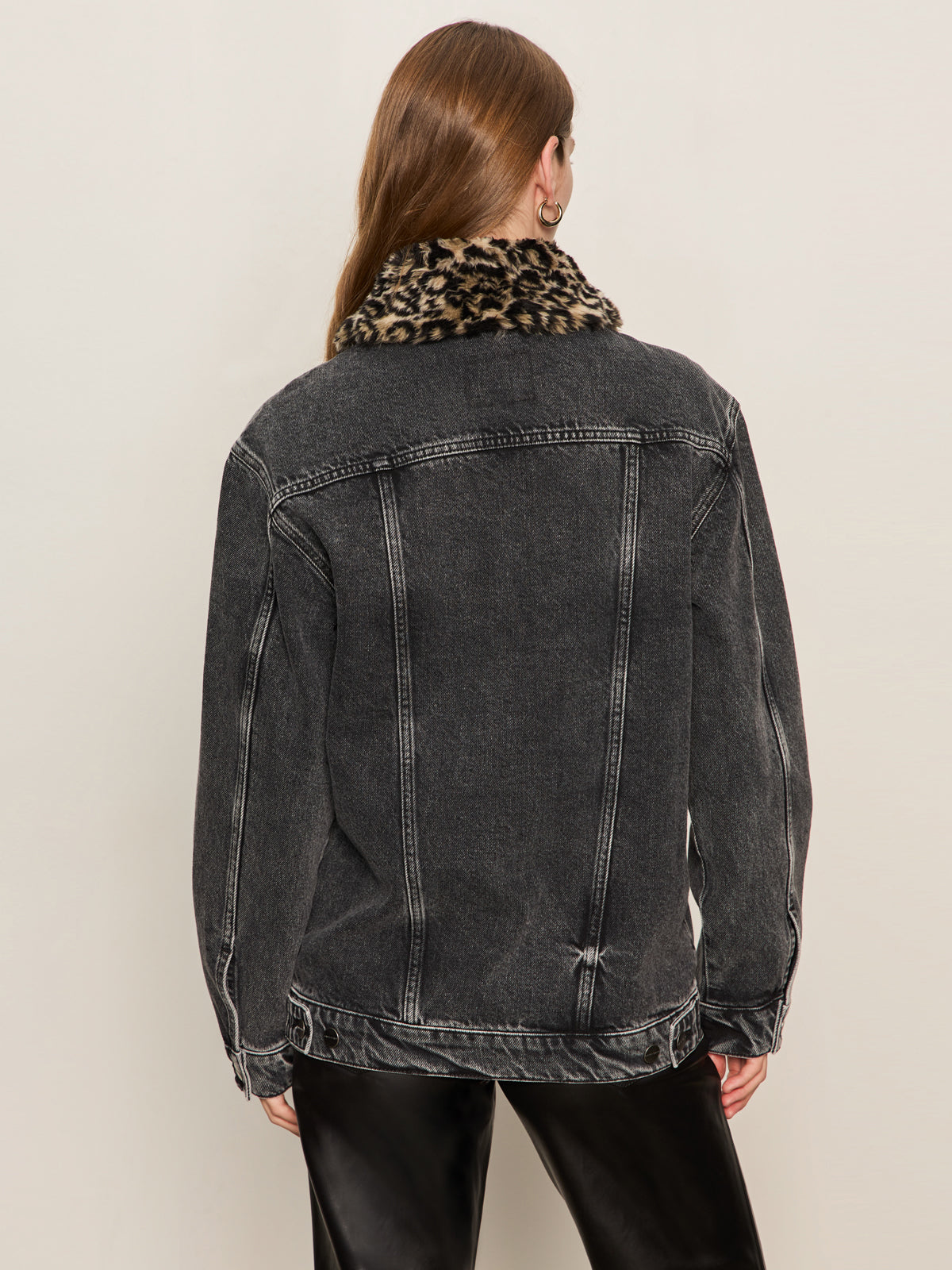 A person with long brown hair is seen from behind wearing the Sanctuary Clothing boyfriend trucker jacket in washed black denim, featuring a leopard print faux fur collar, paired with black pants against a plain light background.