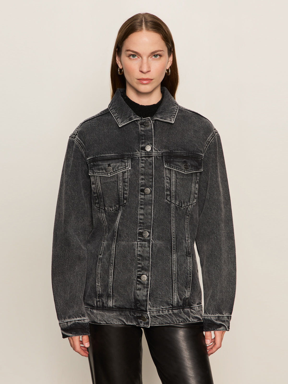 A woman with straight brown hair wears the Sanctuary Clothing boyfriend trucker jacket in washed black denim over a black top and black leather pants, standing against a plain light background, looking directly at the camera.