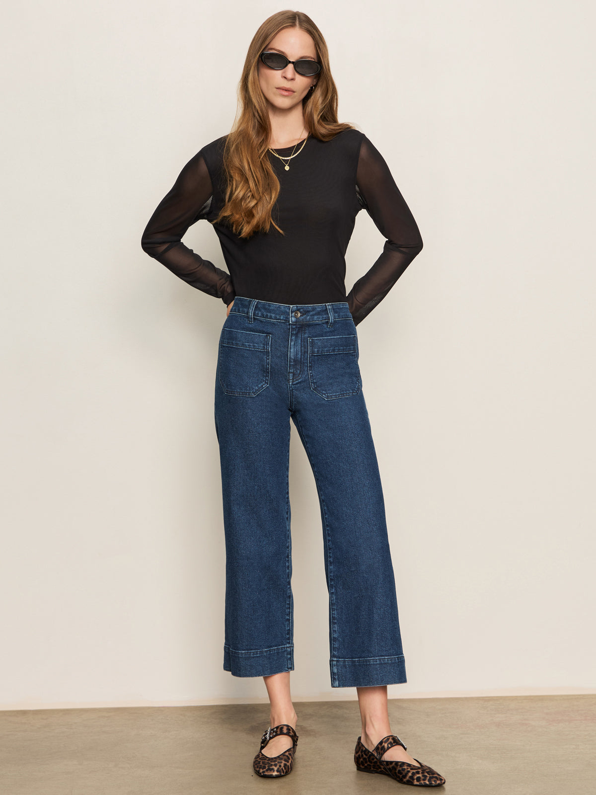 A woman with long, wavy hair stands against a plain background wearing Sanctuary Clothing's the marine jeans aspen, paired with a sheer black top over a black tank, sunglasses, and leopard-print flats. Her hands rest on her hips.