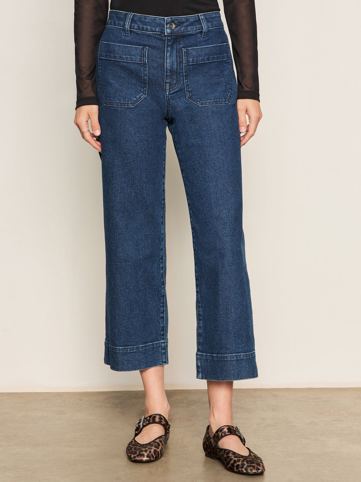 A person wearing Sanctuary Clothing's the marine jeans aspen—high-waisted, cropped blue jeans with front patch pockets—paired with a sheer black long-sleeve top and brown leopard-print flats stands against a neutral background.