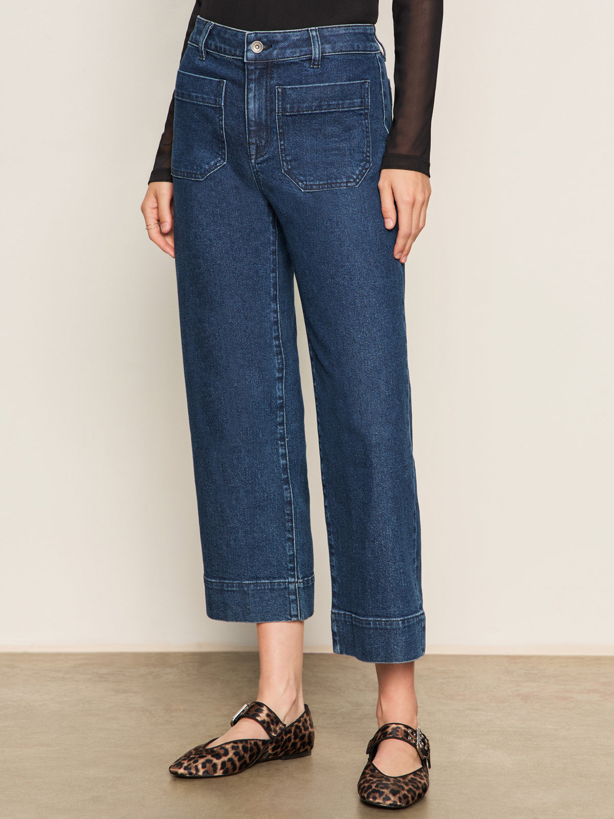 Wearing Sanctuary Clothing's the marine jeans aspen—high-waisted, cropped blue jeans with front patch pockets—a person pairs them with a black long-sleeve top and leopard print Mary Jane flats against a plain background.