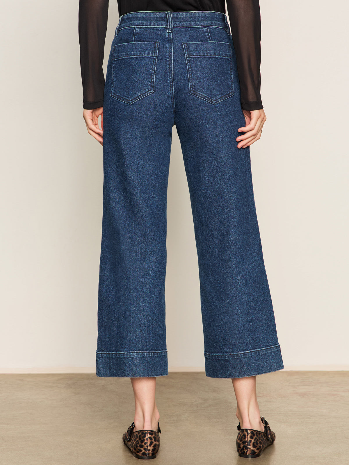 A person wears Sanctuary Clothing's The Marine Jeans Aspen—high-waisted, wide-leg blue jeans with back pockets—paired with a sheer black long-sleeve top and leopard-print flats, standing on a neutral floor facing away.