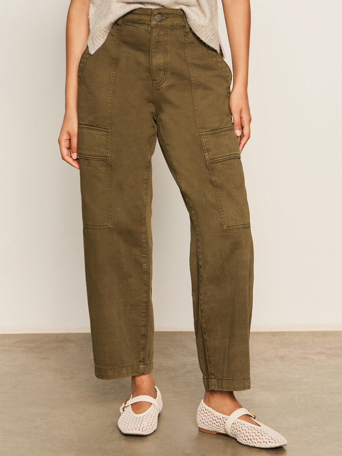 A person wearing Sanctuary Clothing’s boyfriend utility pant kalamata and a light beige top with white woven flats stands on a polished concrete floor against a plain light-colored wall.