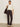 A woman with her hair tied back stands against a plain background wearing Sanctuary Clothing’s Corduroy Hayden Bootcut Pant in Semi Sweet Petites, styled with a cream sweater, beige heeled boots, and a large dark brown bag over her shoulder.
