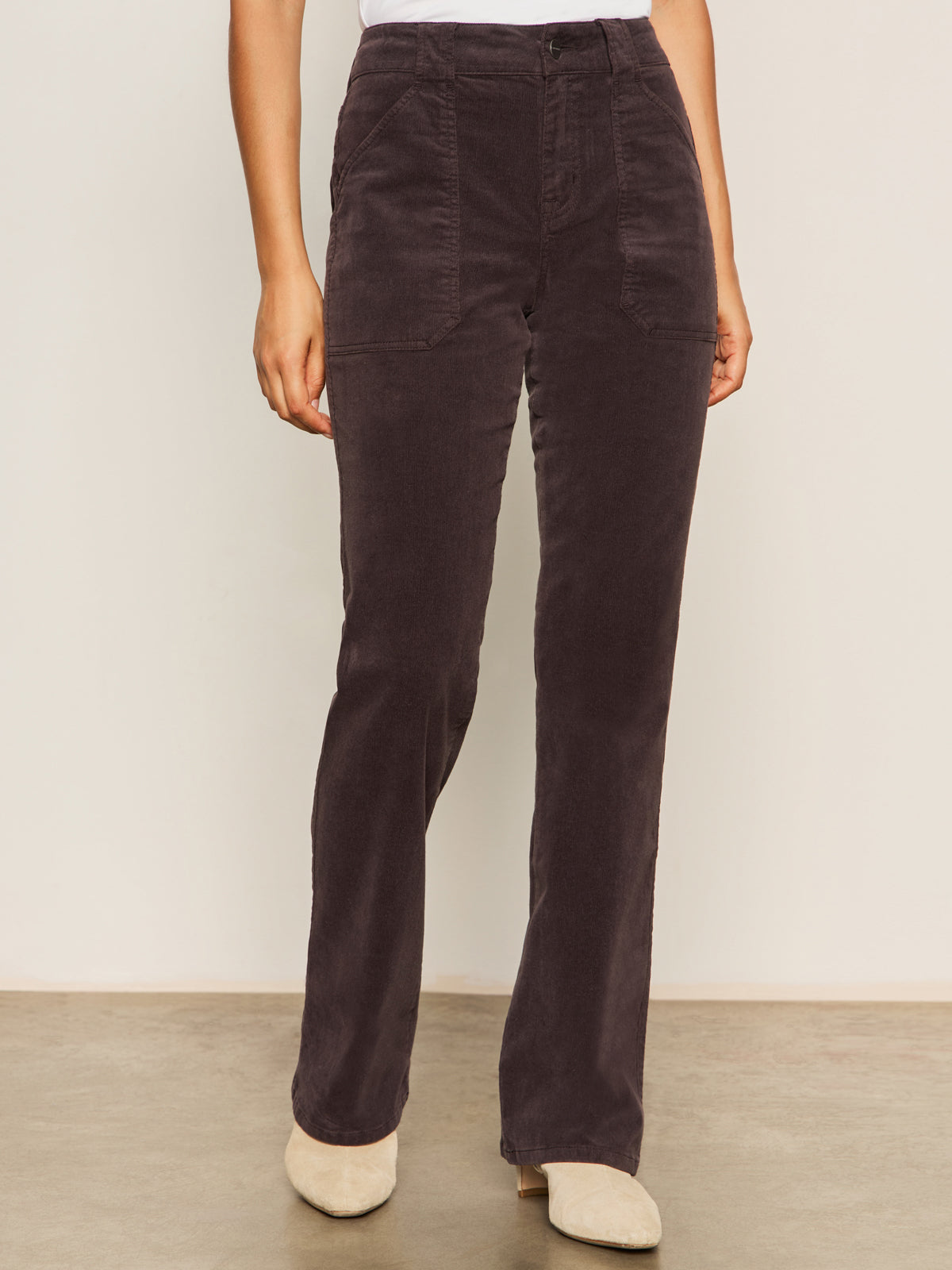 A person wears Sanctuary Clothing's corduroy hayden bootcut pant semi sweet petites in dark brown with front patch pockets, paired with a white top and light beige shoes, standing against a plain background.
