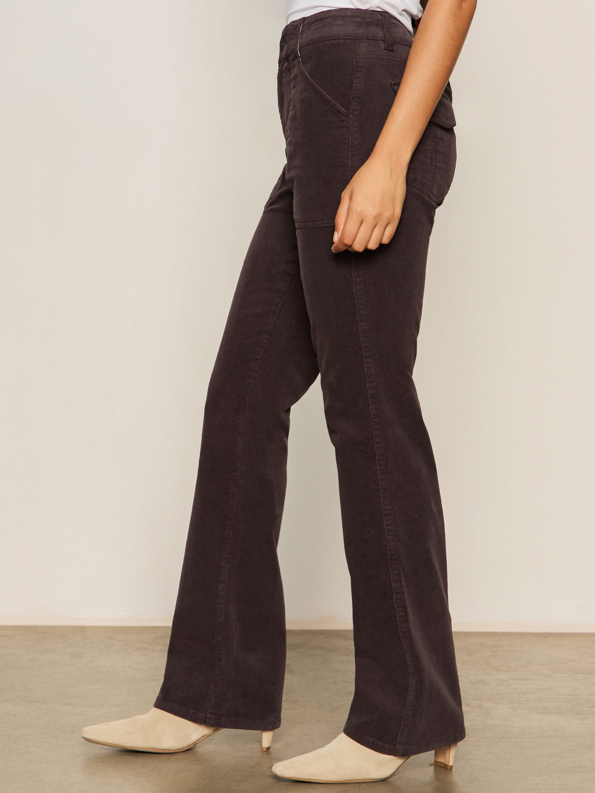 A person in Sanctuary Clothing's corduroy hayden bootcut pant semi sweet petites and light beige heeled boots stands sideways against a plain light-colored wall.