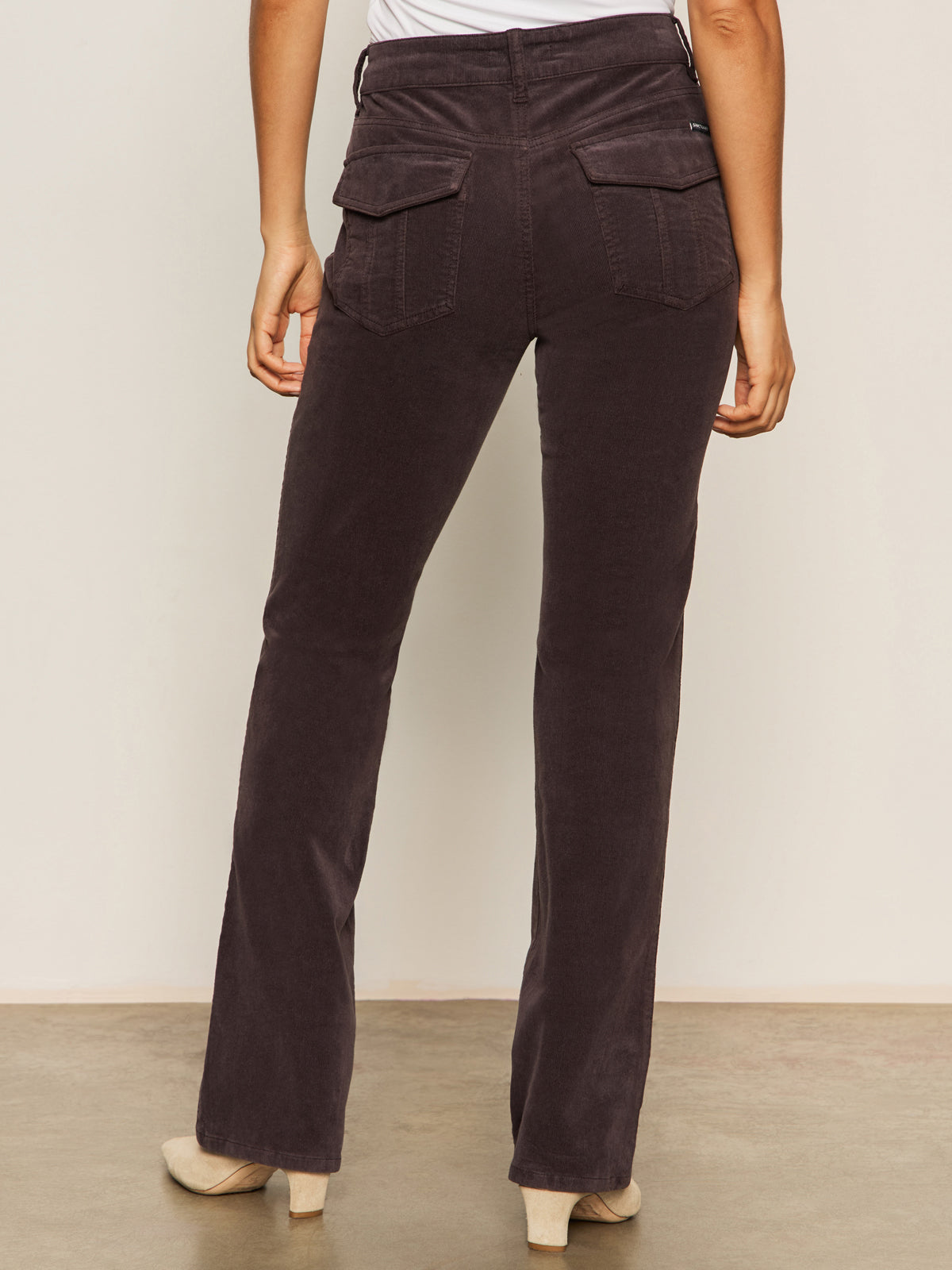 A person stands facing away, wearing Sanctuary Clothing's corduroy hayden bootcut pant semi sweet petites, paired with a white top and beige heeled shoes on a light floor against a plain background.