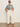 A woman wearing Sanctuary Clothing's "the perfect tee green oasis" (extended sizing), cream wide-leg pants, and sandals stands against a plain off-white wall with her hands in her pockets.