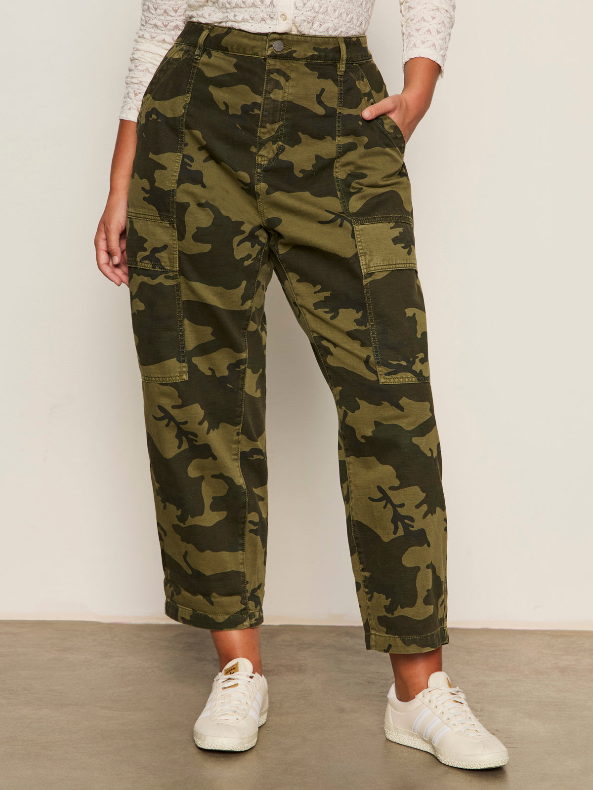 A person stands against a white wall wearing Sanctuary Clothing’s boyfriend utility pant rambler camo (extended sizing), a white patterned blouse, and white sneakers, with one hand in their pants pocket.