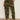 A person stands against a white wall wearing Sanctuary Clothing’s boyfriend utility pant rambler camo (extended sizing), a white patterned blouse, and white sneakers, with one hand in their pants pocket.