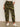 A person stands against a white wall wearing Sanctuary Clothing’s boyfriend utility pant rambler camo (extended sizing), a white patterned blouse, and white sneakers, with one hand in their pants pocket.