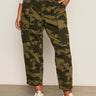 A person stands against a white wall wearing Sanctuary Clothing’s boyfriend utility pant rambler camo (extended sizing), a white patterned blouse, and white sneakers, with one hand in their pants pocket.
