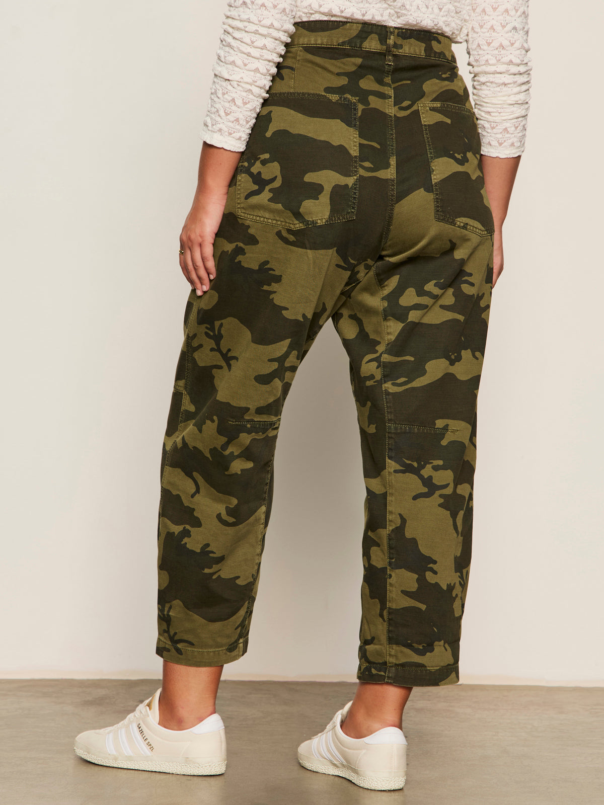 A person faces away, wearing Sanctuary Clothing’s boyfriend utility pant rambler camo (extended sizing)—high-waisted green camo pants with patch pockets and a relaxed fit—paired with white sneakers against a neutral background.