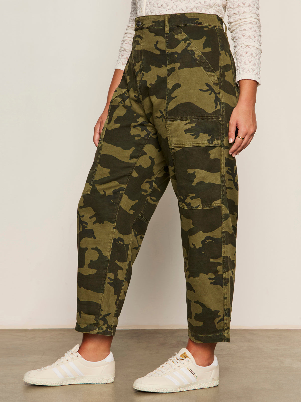 A person stands indoors in Sanctuary Clothing boyfriend utility pant rambler camo (extended sizing), a white textured long-sleeve top, and white Adidas sneakers. The side view photo captures them from shoulders to feet.