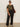 A person wearing sunglasses, a camouflage t-shirt, light shoes, and the Sanctuary Clothing runway ponte trouser black extended sizing stands against a plain wall with a large brown shoulder bag.