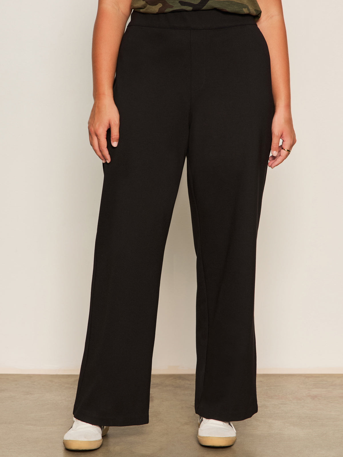 A person wears Sanctuary Clothing's runway ponte trouser in black extended sizing and white sneakers, standing against a plain light-colored wall with their arms relaxed at their sides.