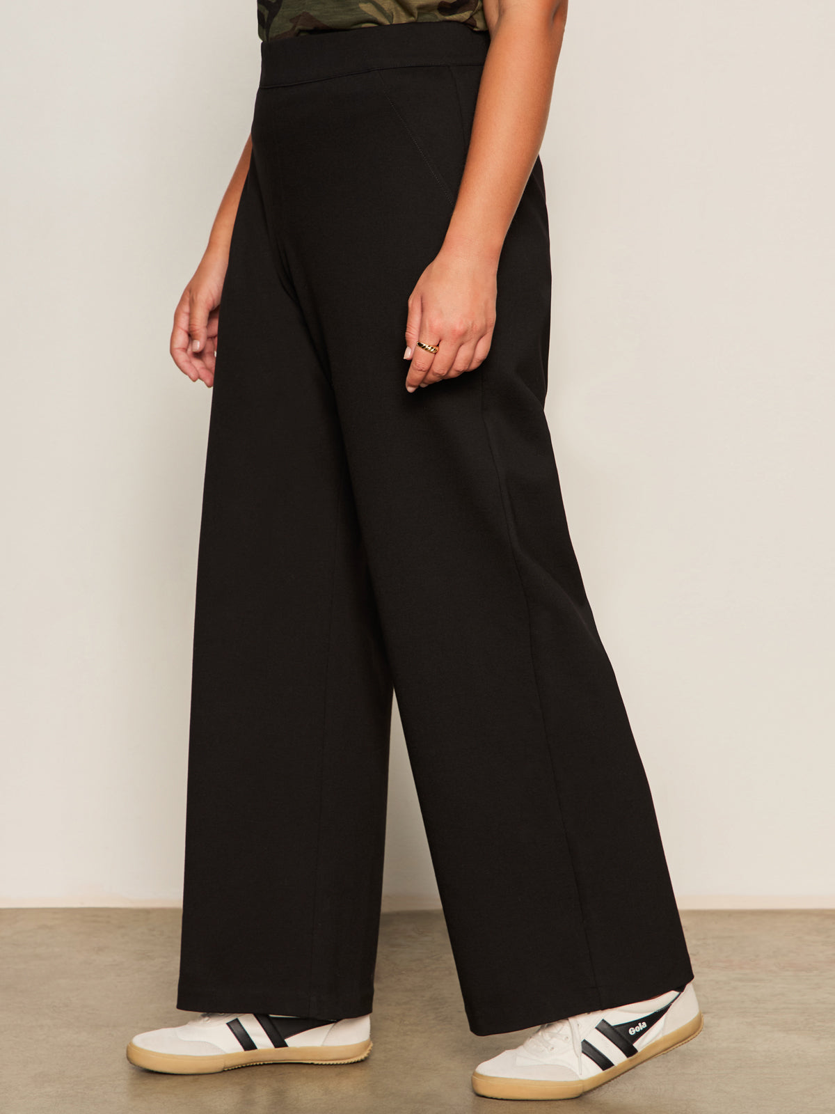 A person in Sanctuary Clothing's runway ponte trouser black (extended sizing), paired with white sneakers featuring a black stripe and a camo-patterned top, poses indoors against a neutral background.