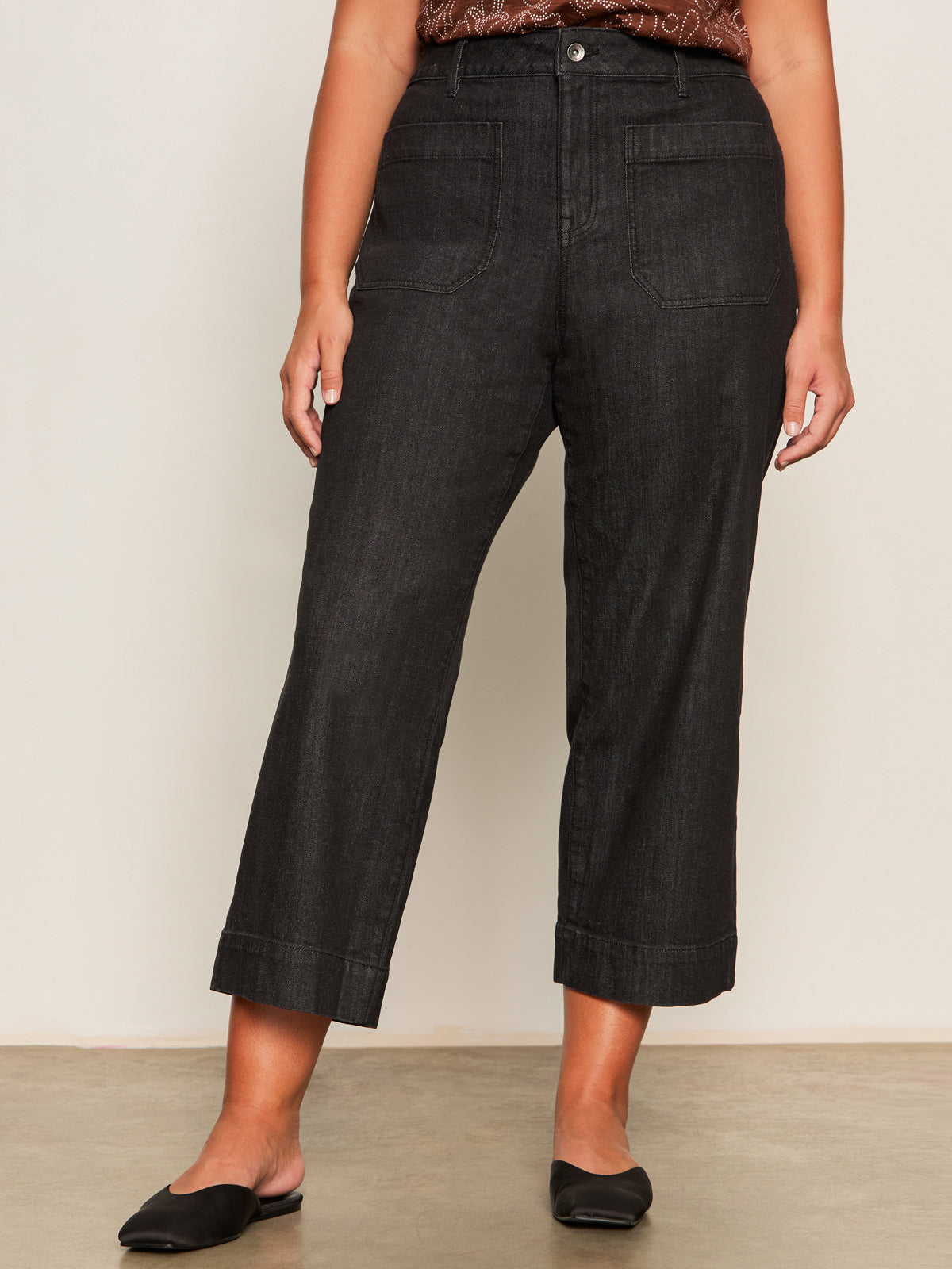 Someone wearing the Marine Jeans After Hours (extended sizing) by Sanctuary Clothing, featuring black cropped wide-leg style with front patch pockets, and black pointed-toe flats, stands against a light wall and concrete floor.