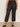Someone wearing the Marine Jeans After Hours (extended sizing) by Sanctuary Clothing, featuring black cropped wide-leg style with front patch pockets, and black pointed-toe flats, stands against a light wall and concrete floor.