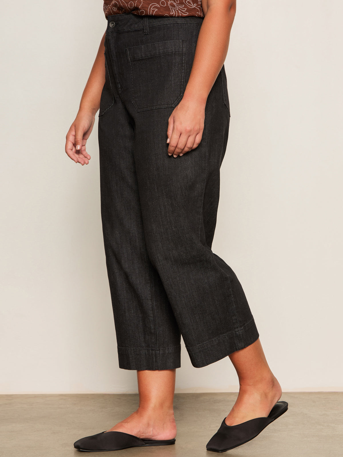 Only the lower half of a person is visible indoors, showcasing Sanctuary Clothing's marine jeans after hours extended sizing—wide-leg, cropped black denim paired with a brown patterned top and black slip-on flats.