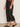 Only the lower half of a person is visible indoors, showcasing Sanctuary Clothing's marine jeans after hours extended sizing—wide-leg, cropped black denim paired with a brown patterned top and black slip-on flats.