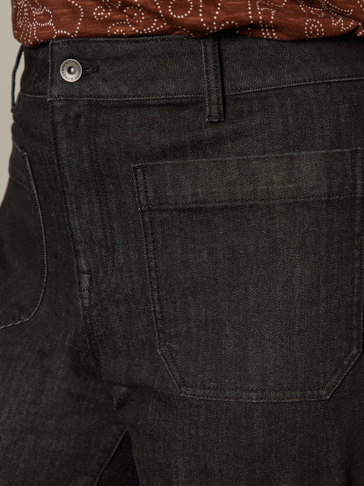 Close-up of a person wearing Sanctuary Clothing’s the marine jeans after hours extended sizing in black denim, highlighting the front pocket and waistband with part of a brown patterned shirt visible at the top.