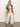 A woman with straight blonde hair poses against a plain background in the Sanctuary Clothing "sanc souvenir shirt mesa palm," a maroon palm-leaf print short-sleeve shirt layered over a white crop top, cream wide-leg pants, and brown sandals.