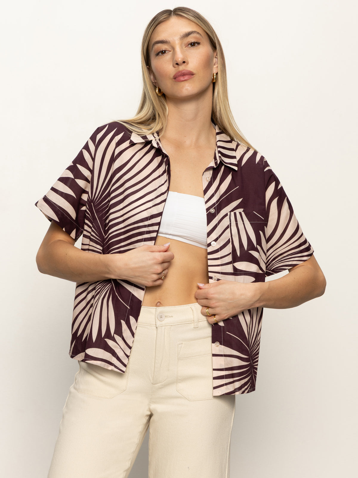 A woman with long blonde hair models the Sanctuary Clothing sanc souvenir shirt mesa palm—a short-sleeve brown shirt with a beige palm leaf design—layered over a white crop top and high-waisted cream pants against a light backdrop.