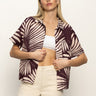 A woman with long blonde hair models the Sanctuary Clothing sanc souvenir shirt mesa palm—a short-sleeve brown shirt with a beige palm leaf design—layered over a white crop top and high-waisted cream pants against a light backdrop.