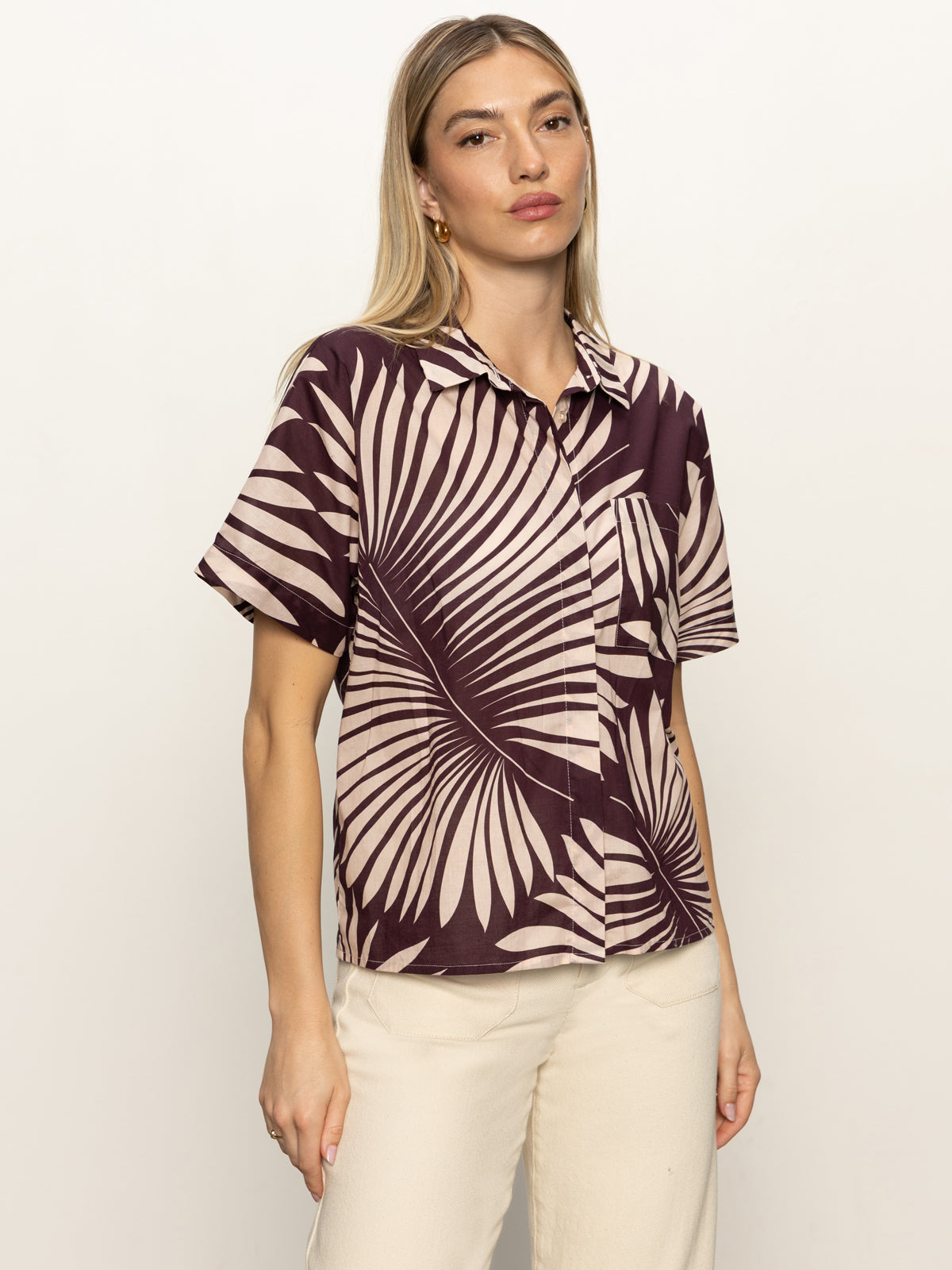 A woman with long blonde hair wears the Sanctuary Clothing sanc souvenir shirt mesa palm—a short-sleeved, button-up shirt with a bold beige palm leaf pattern on dark brown—paired with light pants against a plain backdrop.