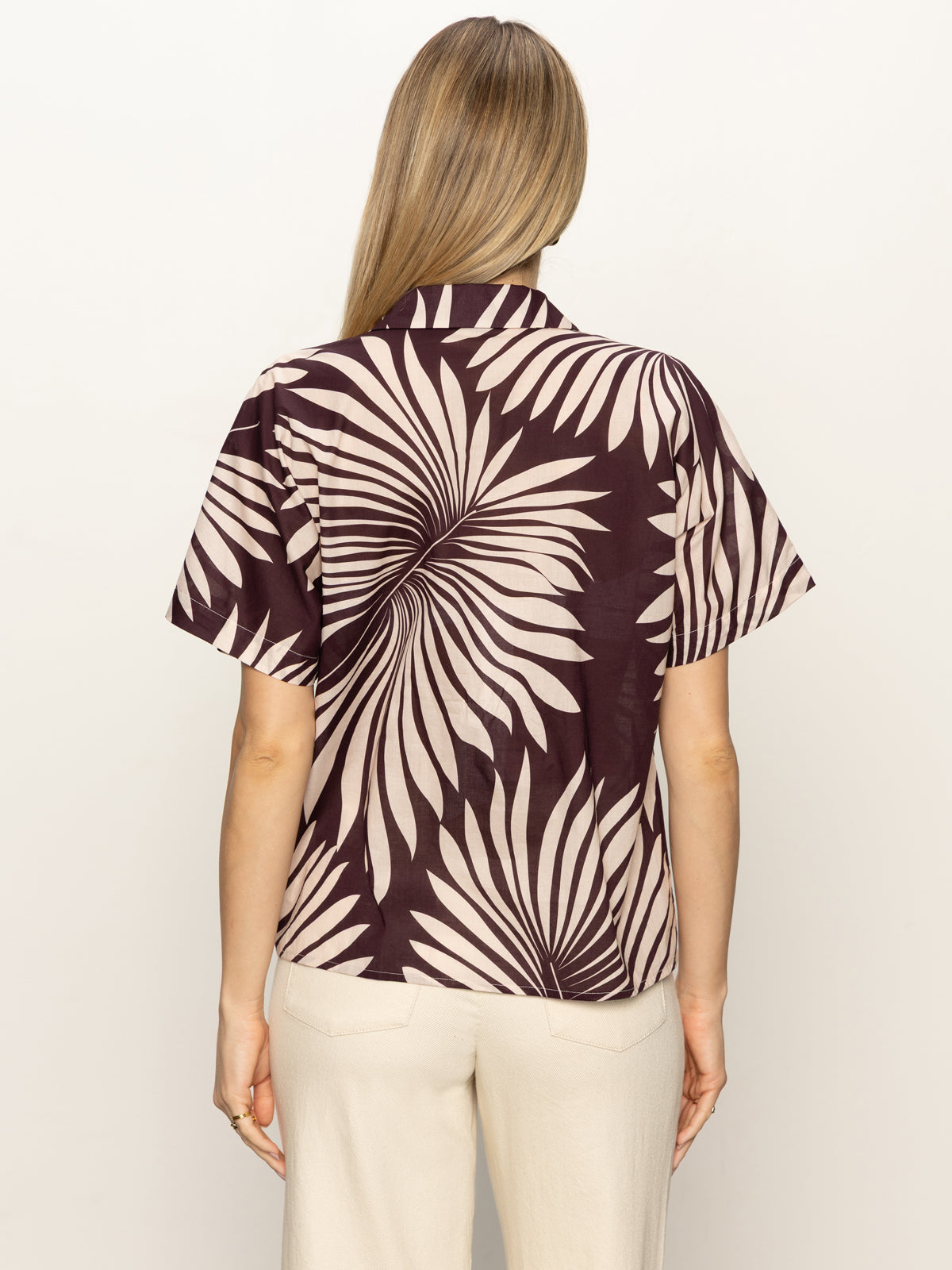 A person with long, straight blonde hair is seen from the back wearing the Sanctuary Clothing sanc souvenir shirt mesa palm—a short-sleeve dark shirt with a large light leaf pattern—paired with light beige pants against a plain background.