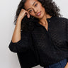 A woman with curly hair sits on a black chair, softly gazing at the camera. She wears blue jeans and the Sanctuary Clothing petal drop blouse in black.